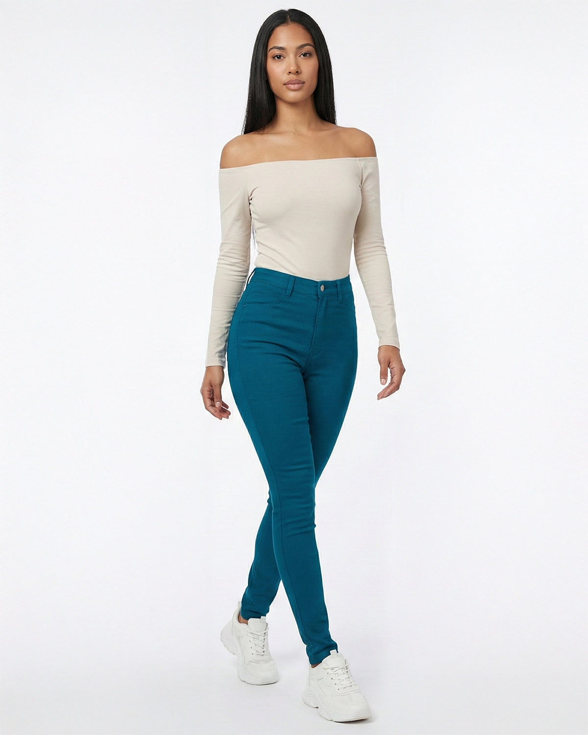 Woman wearing deep teal skinny pants and a beige off-shoulder top on a white background