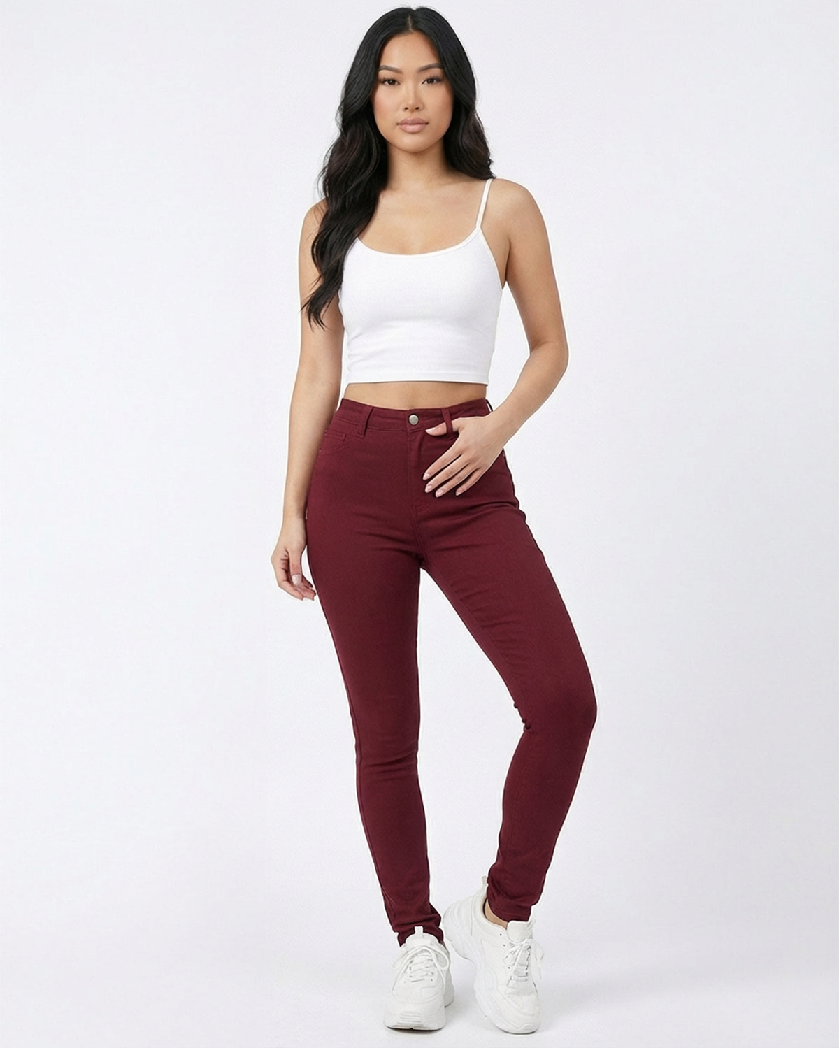 Woman wearing maroon skinny jeans and a white tank top on a white background